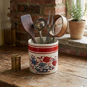 May include: A ceramic kitchen utensil holder with a red stripe design and floral pattern, holding a wooden spatula, metal ladle, fork, and sieve. A small brass container sits to the left. The items are on a wooden surface.