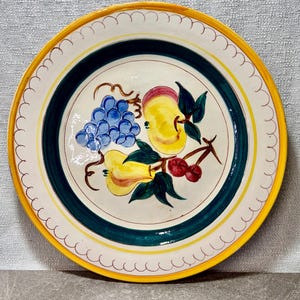 May include: A round, hand-painted ceramic plate with a yellow scalloped edge and a green and yellow border. The center features a still life of fruit, including grapes, pears, cherries, and an apple. The plate is approximately 10 inches in diameter.