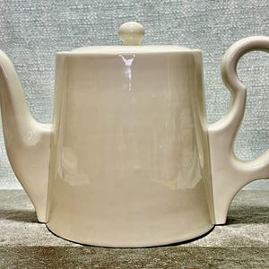 May include: Cream-colored ceramic teapot with a lid and a curved handle. The teapot has a spout and a rounded body. The handle is a unique, open design. The teapot is likely for serving tea.