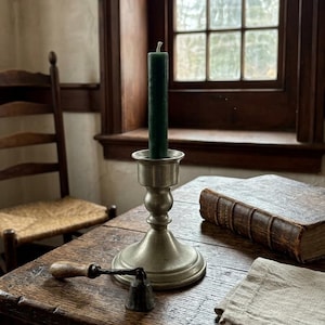 May include: A pewter candlestick holds a dark green candle on a wooden table. A book, a candle snuffer, and a linen cloth are also on the table. A wooden chair and a window are in the background.