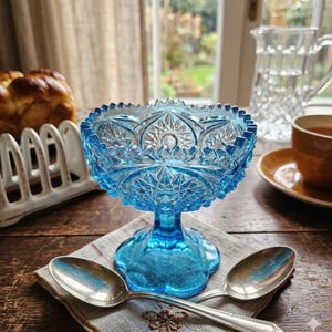 May include: A vibrant blue glass pedestal bowl with intricate starburst and geometric patterns. Two silver spoons rest on a linen napkin, alongside a cup of tea and a bread basket. The bowl is approximately 6 inches tall.