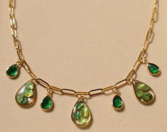 Puede incluir: Un collar de cadena en tono dorado con una serie de dijes colgantes. Los dijes alternan entre formas de gota de color verde esmeralda sólido y colgantes transparentes en forma de gota que contienen pequeños fragmentos de concha iridiscentes.