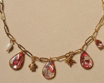 Puede incluir: Un collar de cadena dorada con varios dijes, incluyendo dos colgantes en forma de lágrima rellenos de fragmentos de conchas rosas y blancas, un dije de tortuga, una estrella, una perla blanca y una gota de cristal rosa.