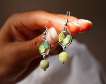 Puede incluir: Un par de pendientes colgantes con un marco circular de tono plateado que contiene pequeñas piedras lisas de color verde claro y amarillo. Debajo de cada marco circular cuelga una cuenta redonda de color verde pálido sujeta por un pequeño eslabón metálico.