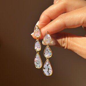 Pode incluir: Um par de brincos pendentes com três pingentes em forma de gota alinhados verticalmente. Cada pingente é envolto numa moldura de metal dourado e preenchido com fragmentos de concha iridescentes e multicoloridos que refletem tons de rosa, azul e branco.