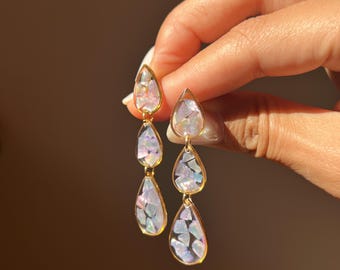 Puede incluir: Un par de pendientes colgantes con tres dijes en forma de lágrima alineados verticalmente. Cada dije está engastado en un marco de metal dorado y relleno con fragmentos de concha iridiscentes y multicolores que reflejan tonos rosas, azules y blancos.