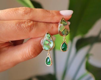 Puede incluir: Un par de pendientes colgantes con un marco dorado en forma de lágrima relleno de resina transparente e incrustaciones de fragmentos de concha verde iridiscente, con un pequeño dije de cristal verde en forma de lágrima colgando.