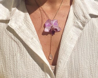 Puede incluir: Un collar dorado con un colgante con una flor de orquídea de color púrpura claro y una piedra en forma de lágrima. El collar se lleva sobre una camisa blanca texturizada.