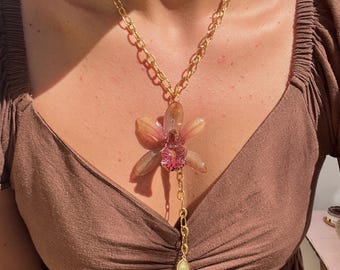 Puede incluir: Un collar de cadena dorada con un colgante grande con diseño de flor. La flor tiene una combinación de colores rosa claro y beige con un centro rosa más pequeño. Una cadena dorada más pequeña con un dije en forma de lágrima cuelga debajo.