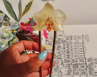 Puede incluir: Una horquilla de metal dorado con una flor de orquídea de color crema translúcido preservada en la parte superior. La flor tiene un detalle central naranja y rojo oscuro, y la horquilla de dos puntas tiene un acabado liso y pulido.