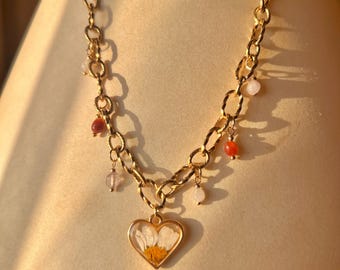 Puede incluir: Un collar de cadena en tono dorado con un colgante en forma de corazón que contiene una flor de margarita blanca prensada con el centro amarillo. La cadena está adornada con pequeñas cuentas de piedras preciosas multicolores en tonos rojo, naranja y rosa pálido.
