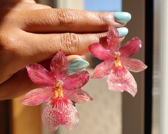 Puede incluir: Un par de pendientes con flores de orquídea preservadas en tonos rosa intenso y magenta. Cada flor tiene un acabado translúcido y brillante con vetas intrincadas y un patrón moteado en los pétalos inferiores, mostrando detalles botánicos naturales.