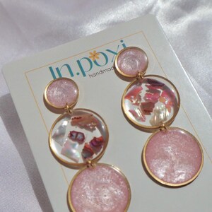 Pode incluir: Brincos rosa e dourados feitos à mão com um design em camadas. Os brincos apresentam três elementos circulares, com os círculos inferior e superior em cor rosa sólida e o círculo do meio preenchido com peças iridescentes. Os brincos estão num cartão branco com o texto "in.poxy handmade".