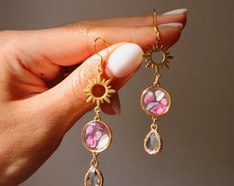 Puede incluir: Pendientes colgantes dorados con un dije en forma de sol conectado a un colgante circular relleno de fragmentos de piedra rosa y blanca, rematados con un cristal transparente en forma de lágrima que cuelga de un gancho dorado.