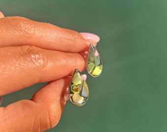 Puede incluir: Un par de pendientes de botón en forma de lágrima con un marco de metal plateado relleno de un mosaico de fragmentos de concha iridiscentes y multicolores en tonos verde, amarillo y rosa, sobre una base de resina oscura translúcida.