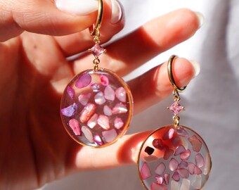 Puede incluir: Un par de pendientes colgantes circulares con bordes y ganchos de metal dorado. Cada pendiente contiene fragmentos de piedra rosa y morada incrustados en un centro de resina transparente, con un pequeño cristal rosa de corte cuadrado.