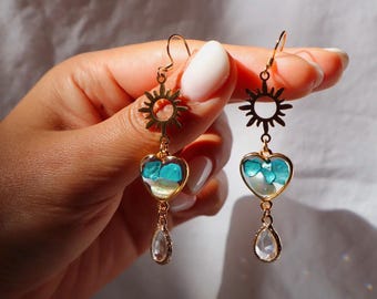 Puede incluir: Un par de pendientes colgantes de tono dorado con un dije en forma de sol en la parte superior, un colgante en forma de corazón relleno de pequeñas piedras turquesas y blancas, y un cristal transparente en forma de lágrima bajo cada corazón.
