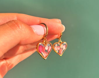 Puede incluir: Un par de pendientes de aro dorados con colgantes en forma de corazón. Cada colgante tiene un marco dorado que encierra un centro de resina transparente relleno de pequeños fragmentos de piedra de colores rosa y blanco.