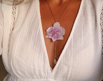 Puede incluir: Un collar tipo lariat en tono dorado con un colgante de flor de orquídea translúcida en tonos blanco y violeta pálido. Una delicada cadena dorada desciende desde el centro de la orquídea y termina en un pequeño dije de piedra iridiscente en forma de gota.