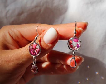 Puede incluir: Un par de pendientes colgantes de tono plateado con un dije circular relleno de pequeños fragmentos de piedra rosa y roja, suspendido sobre una cuenta de cristal transparente en forma de lágrima sujeta a un gancho estándar.
