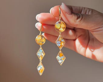 Puede incluir: Un par de pendientes colgantes dorados con un dije circular superior y dos dijes en forma de diamante suspendidos debajo. Cada dije está relleno de resina transparente que contiene pequeños fragmentos de piedra color ámbar y azul claro.