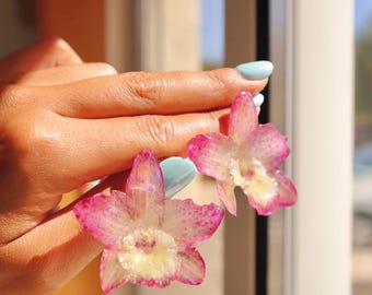 Puede incluir: Un par de pendientes florales translúcidos con delicados pétalos moteados en rosa y magenta con un centro amarillo pálido. Los adornos en forma de flor tienen un acabado brillante similar a la resina que refleja la luz del sol.