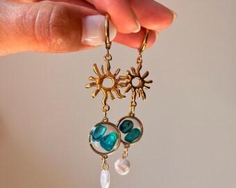 Puede incluir: Un par de pendientes colgantes de tono dorado con un dije en forma de sol conectado a un colgante circular que contiene pequeñas piedras turquesas pulidas. Cada pendiente termina con una pequeña perla blanca de forma irregular colgando.