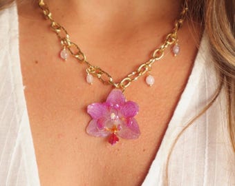 Puede incluir: Collar de cadena dorada con un gran colgante de orquídea rosa translúcida. El collar tiene pequeños detalles de cuentas rosas y se lleva sobre una camisa blanca.
