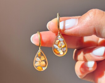 Puede incluir: Un par de pendientes de aro dorados con colgantes en forma de lágrima. Cada colgante está hecho de resina transparente con pequeños fragmentos de piedra pulida incrustados en tonos ámbar, naranja y blanco, suspendidos de un aro dorado fino.