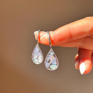 Può includere: Un paio di orecchini a goccia con una cornice in resina trasparente riempita di frammenti di conchiglia iridescenti e multicolori. Ogni pendente è appeso a un semplice cerchio in metallo argentato, riflettendo la luce con un riflesso madreperlaceo.