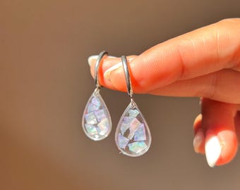 Puede incluir: Un par de pendientes en forma de lágrima con un marco de resina transparente relleno de fragmentos de concha iridiscentes y multicolores. Cada colgante cuelga de un aro de metal plateado, reflejando la luz con un acabado nacarado.