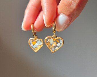 Puede incluir: Un par de pendientes de aro dorados con colgantes en forma de corazón. Cada colgante tiene un marco de metal dorado relleno de resina transparente con pequeños fragmentos de piedra de colores en tonos blanco, gris y naranja.