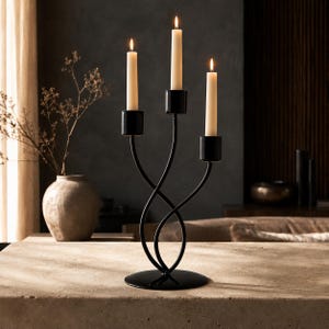 May include: A black metal candelabra with a circular base and three curved arms holding three lit off-white taper candles. The structure features a symmetrical design with smooth, slender metal rods that twist upward to support the candle cups.