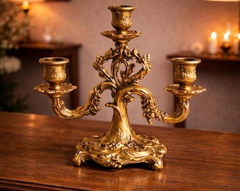 May include: A gold-colored metal candelabra featuring three candle holders arranged in a tiered formation. The base and arms are decorated with intricate, raised floral and leaf motifs, with a central stem supporting the tallest holder.