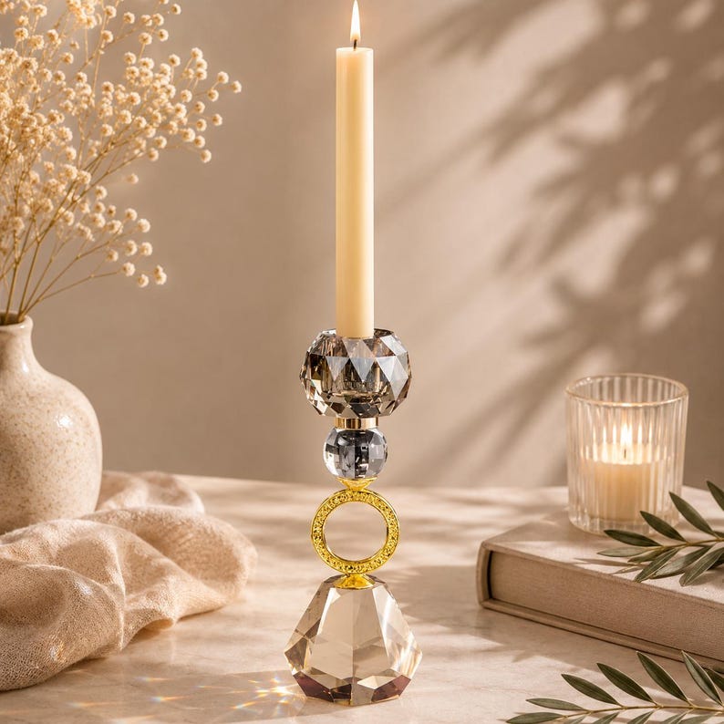 May include: A tall, elegant candle holder with a lit cream-colored candle. The holder features a faceted crystal base, a gold ring, and crystal spheres. A small glass candle holder with a lit candle is in the background.