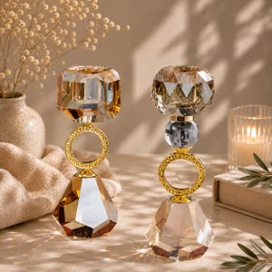 May include: Two decorative candle holders with faceted crystal designs in amber and clear tones. Each holder features a gold-colored ring and base, with a geometric shape. The candle holders are designed to hold a single candle.