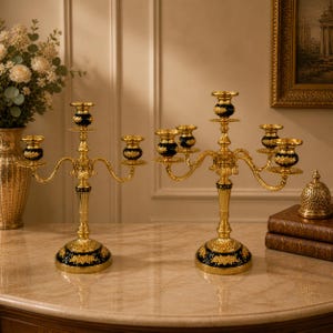 Könnte beinhalten: Zwei goldene Kerzenständer mit schwarzen und goldenen floralen Akzenten. Jeder Kerzenständer hat mehrere Arme für Kerzen und steht auf einem runden Sockel. Die Kerzenständer stehen auf einer marmorähnlichen Oberfläche. Höhe ca. 30 cm.