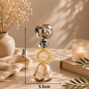 May include: A decorative crystal candle holder with a faceted base and top, connected by a gold ring and a gray crystal sphere. The candle holder is 6.7 inches tall and 2.2 inches wide. The candle holder is designed to hold a single candle.