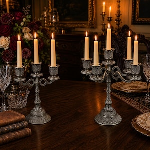 Può includere: Due candelabri in metallo color argento decorati con intricati motivi in rilievo. Il candelabro più piccolo sostiene tre candele affusolate color crema, mentre quello più grande ne sostiene cinque, tutte accese con fiamme visibili.