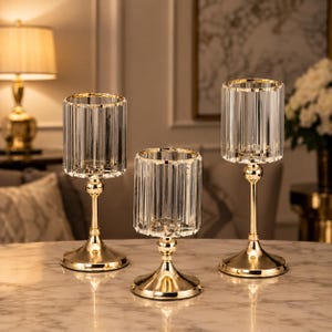 May include: Three decorative candle holders with a gold base and a clear, ribbed glass cylinder. The candle holders are of varying heights and are arranged on a marble-effect surface. The gold base and rim of the glass add a touch of elegance.