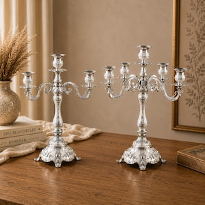 May include: Two silver candelabras with ornate detailing. Each candelabra has multiple arms to hold candles, with a central stem and a decorative base. The design features floral and scrollwork patterns, creating an elegant and classic look.