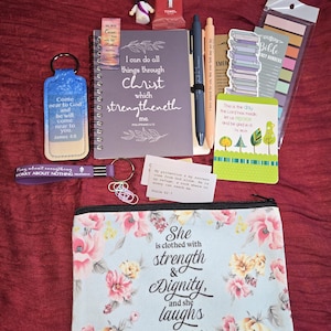 Może przedstawiać: Kolekcja przedmiotów o tematyce religijnej na czerwonej powierzchni. Zawiera dziennik z tekstem, brelok, długopisy, piórnik z kwiatowym wzorem i tekstem, zakładki, małą figurkę i fioletową opaskę na nadgarstek.