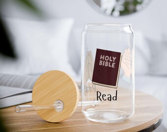 May include: A clear glass can-shaped cup with a bamboo lid and straw. The cup features a graphic of a maroon Holy Bible, praying hands, and the word "Read". The cup is on a wooden table.