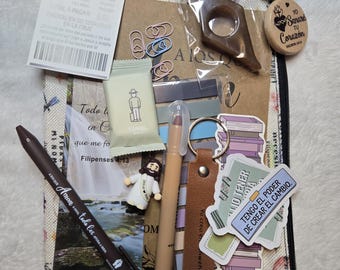 May include: A collection of stationery items, including a pen, stickers, paperclips, and a bookmark, arranged on a patterned surface. The items include a small Jesus figurine, a keychain, and a small notepad with Spanish text.