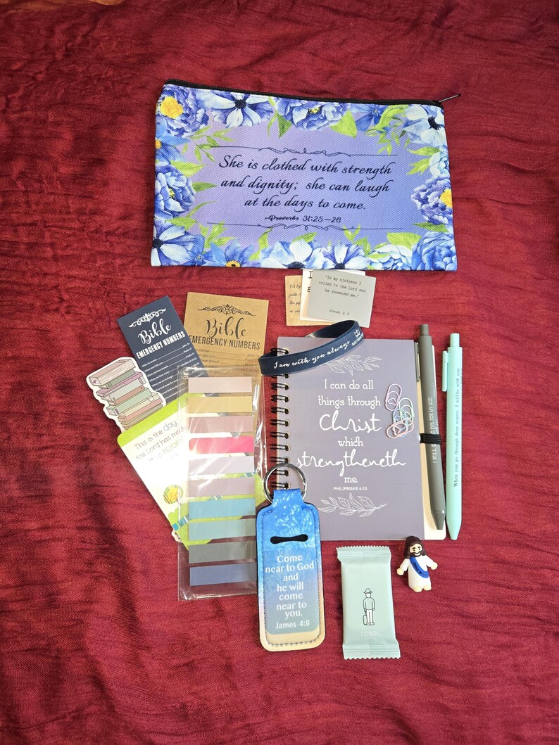 May include: A zippered pouch with blue floral patterns and the text She is clothed with strength and dignity, a spiral notebook with the text I can do all things through Christ, two pens, sticky note tabs, a keychain, and small religious items.