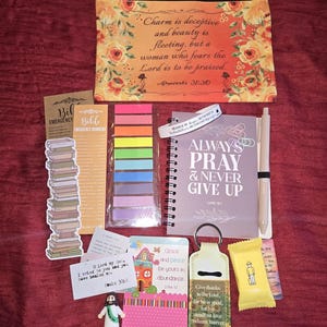 May include: A collection of religious items including a floral pouch with Proverbs 31:30 text, a spiral notebook with Always Pray & Never Give Up text, a pen, colorful sticky tabs, a keychain, a small figurine, and various cards with scripture verses.