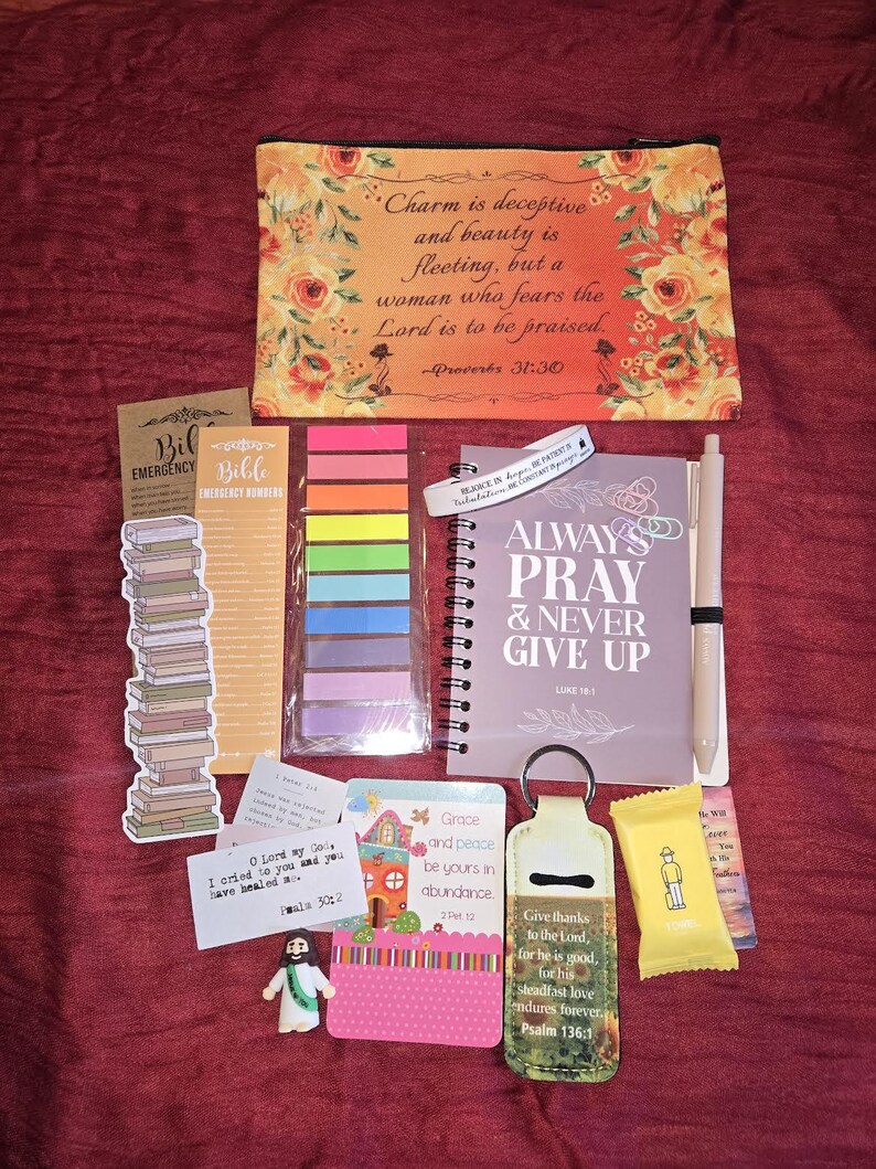 May include: A collection of religious items including a floral pouch with Proverbs 31:30 text, a spiral notebook with Always Pray & Never Give Up text, a pen, colorful sticky tabs, a keychain, a small figurine, and various cards with scripture verses.