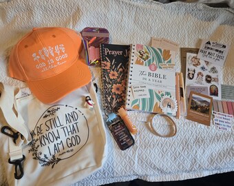 May include: An orange baseball cap with white floral text, a white canvas tote bag with black text, a spiral-bound prayer journal, a Bible in a Year book, a keychain, a lip balm, pens, stickers, and small cards featuring religious themes and quotes.