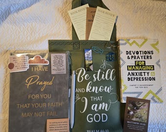 May include: A dark gray journal with gold text, a green tote bag with white text reading Be still and know that I am God, a book titled Devotions & Prayers for Managing Anxiety & Depression, a pen, bookmarks, and paper clips on a white surface.