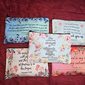May include: Five rectangular zippered pouches featuring floral patterns and printed Bible verses. The pouches display various color schemes including blue, pink, and white, with black text detailing scriptures such as Proverbs 31:25 and 31:26.